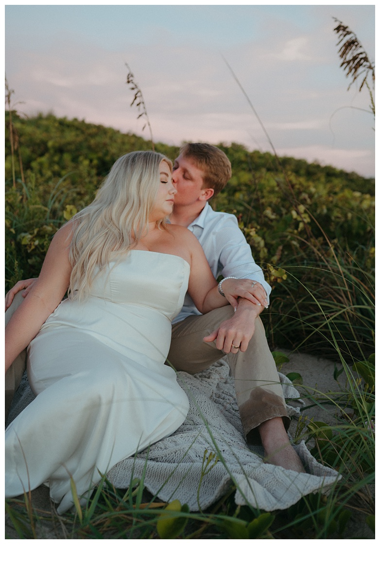 groom and bride sitting within berm while groom kisses brides temple after florida beach elopement