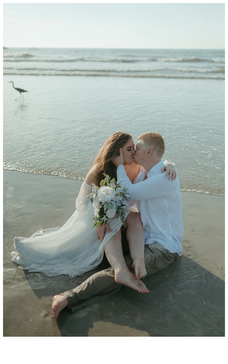 bride and groom sitting on sand kissing