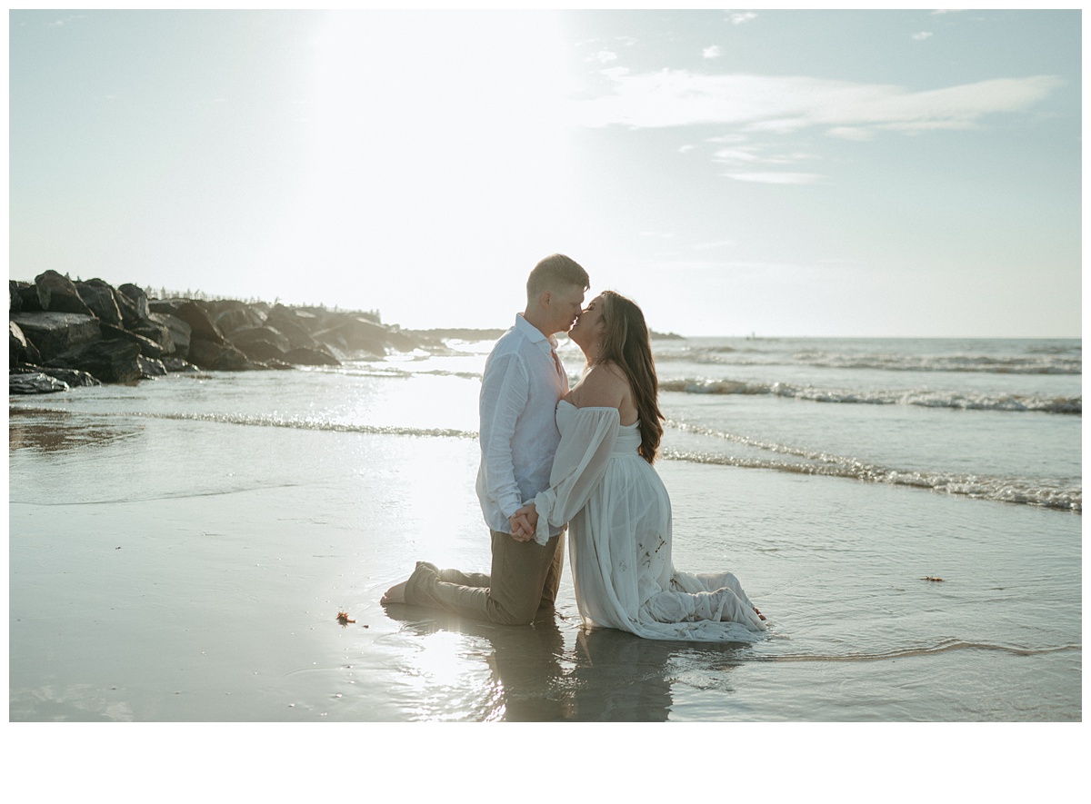 bride and groom holding hands on knees while kissing on beach