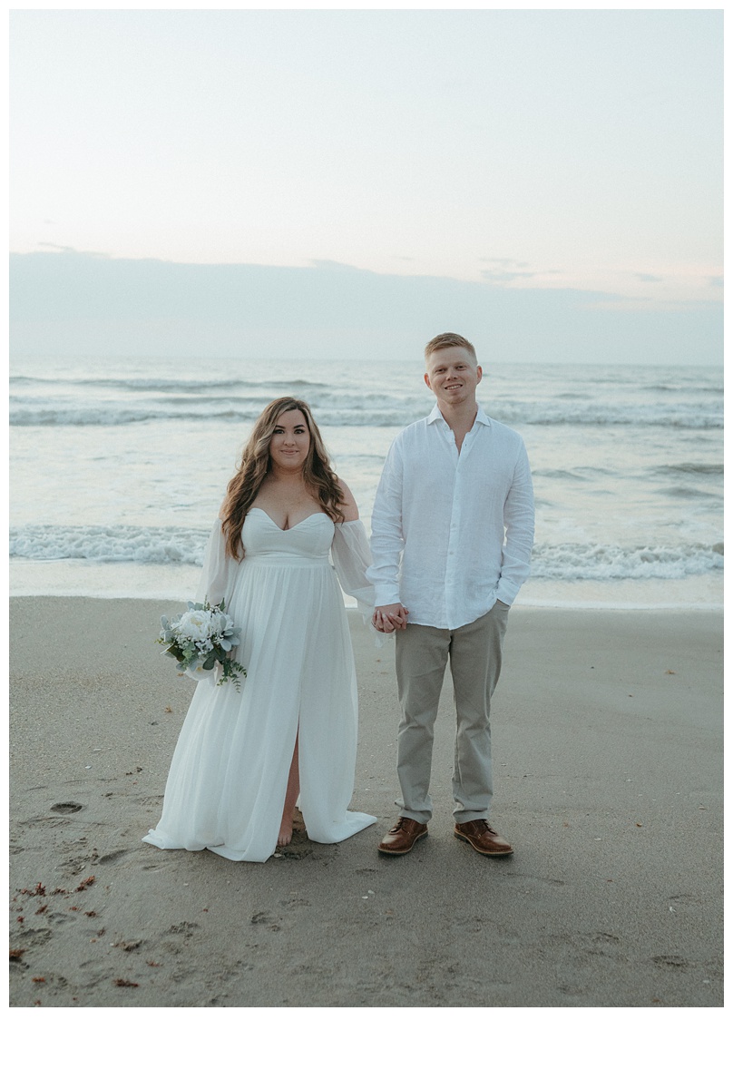 moody bride and groom looking at camera holding hands on beach