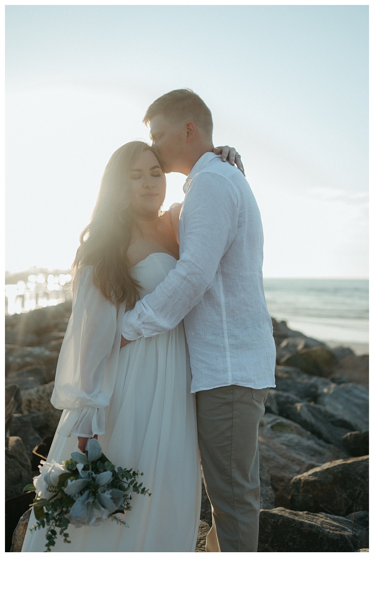 groom kissies brides temple on jetty rocks at beach