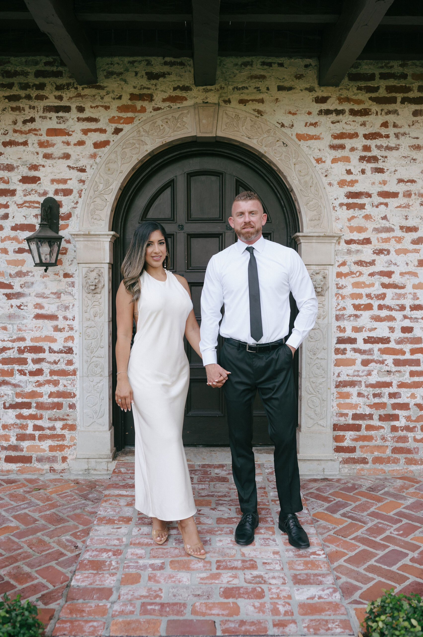 girl wearing a white dress holding hands with a guy wearing a white shirt black pants and a tie. Standing in front of a door